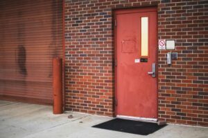A red industrial entrance door on a brick wall exterior, with security access panel and no smoking sign.